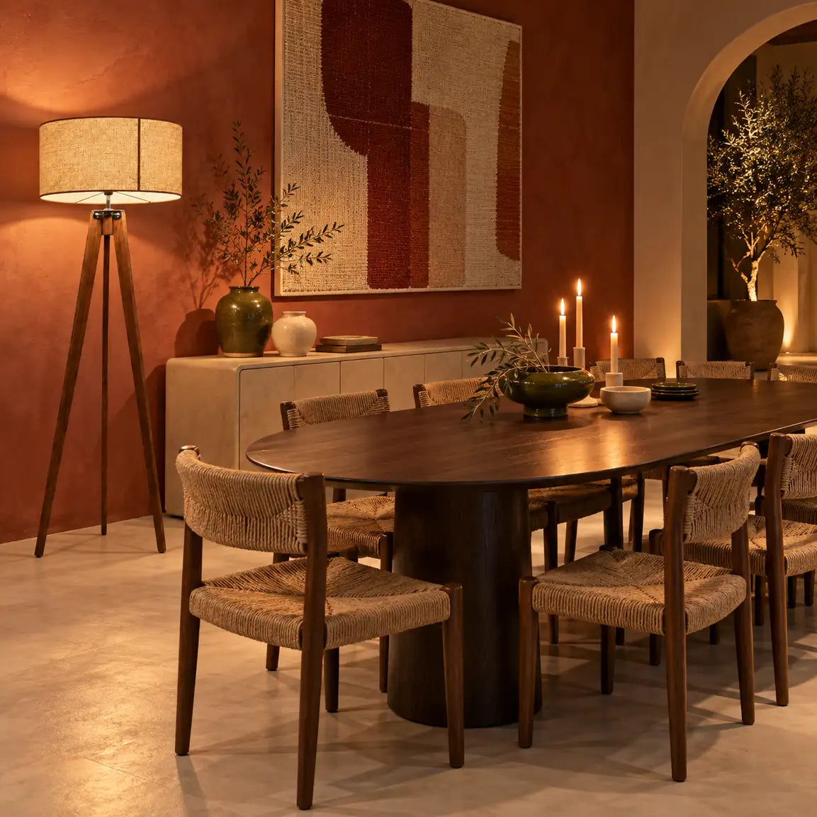 Lampadaire trépied en bois allumé dans une salle à manger terracotta, près d'une table ovale avec bougies et chaises tressées