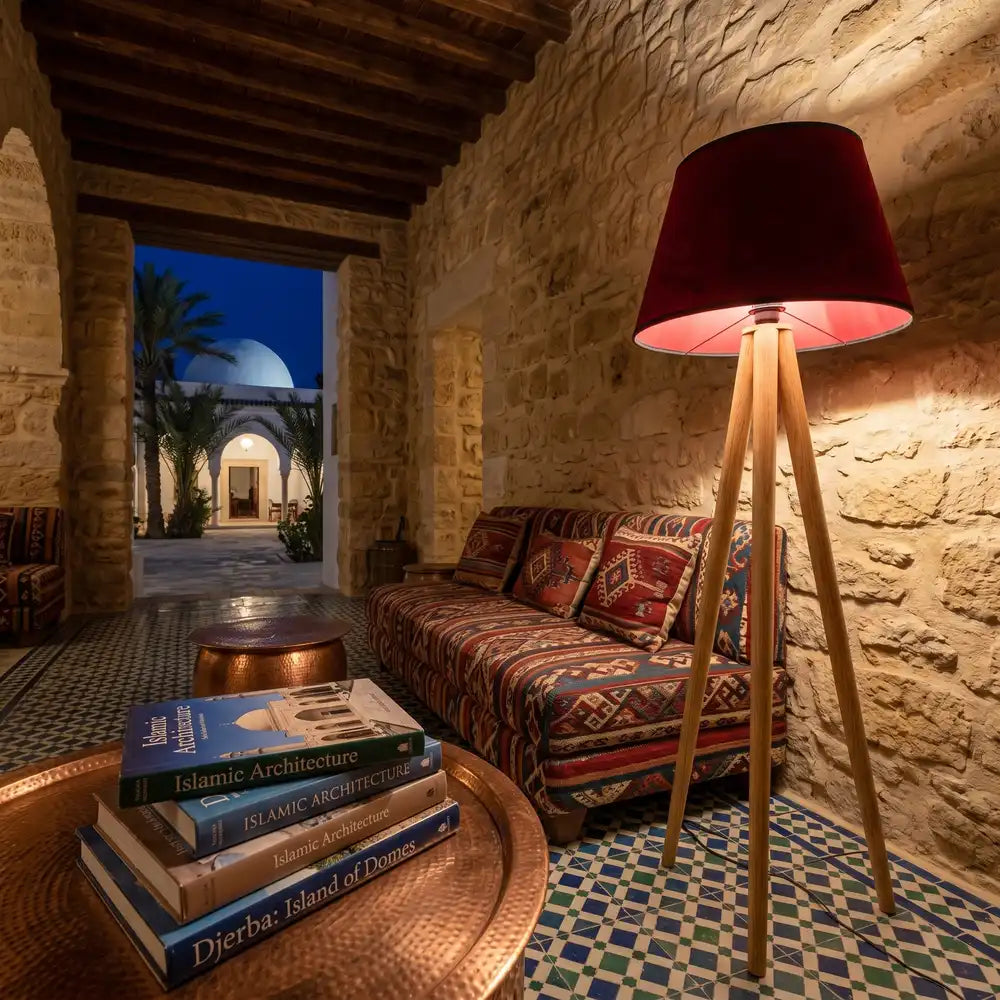 Lampadaire à abat-jour bordeaux allumé le soir près d'une banquette à motifs, dans une pièce en pierre ouvrant sur une cour à coupole blanche