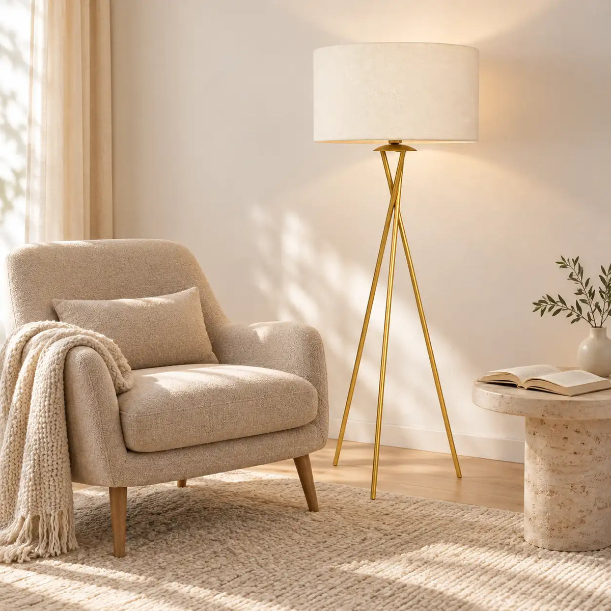 Lampadaire trépied doré dans un coin lecture, entre un fauteuil beige et une table d'appoint en pierre avec livre ouvert