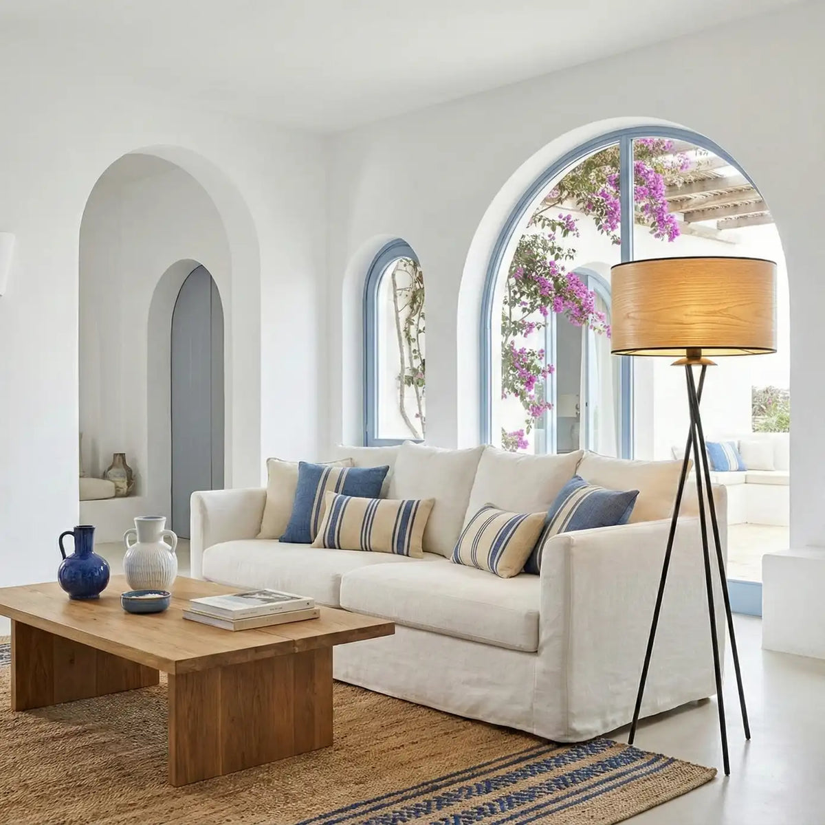 Lampadaire trépied noir à abat-jour effet bois dans un salon blanc méditerranéen aux fenêtres en arche bleues et bougainvilliers