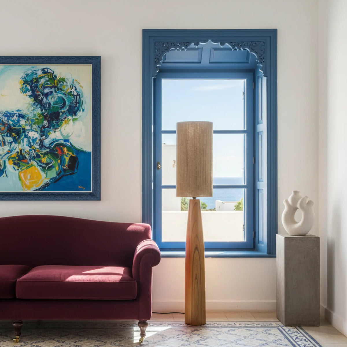 Lampadaire en bois et jute devant une fenêtre à encadrement bleu, à côté d'un canapé bordeaux