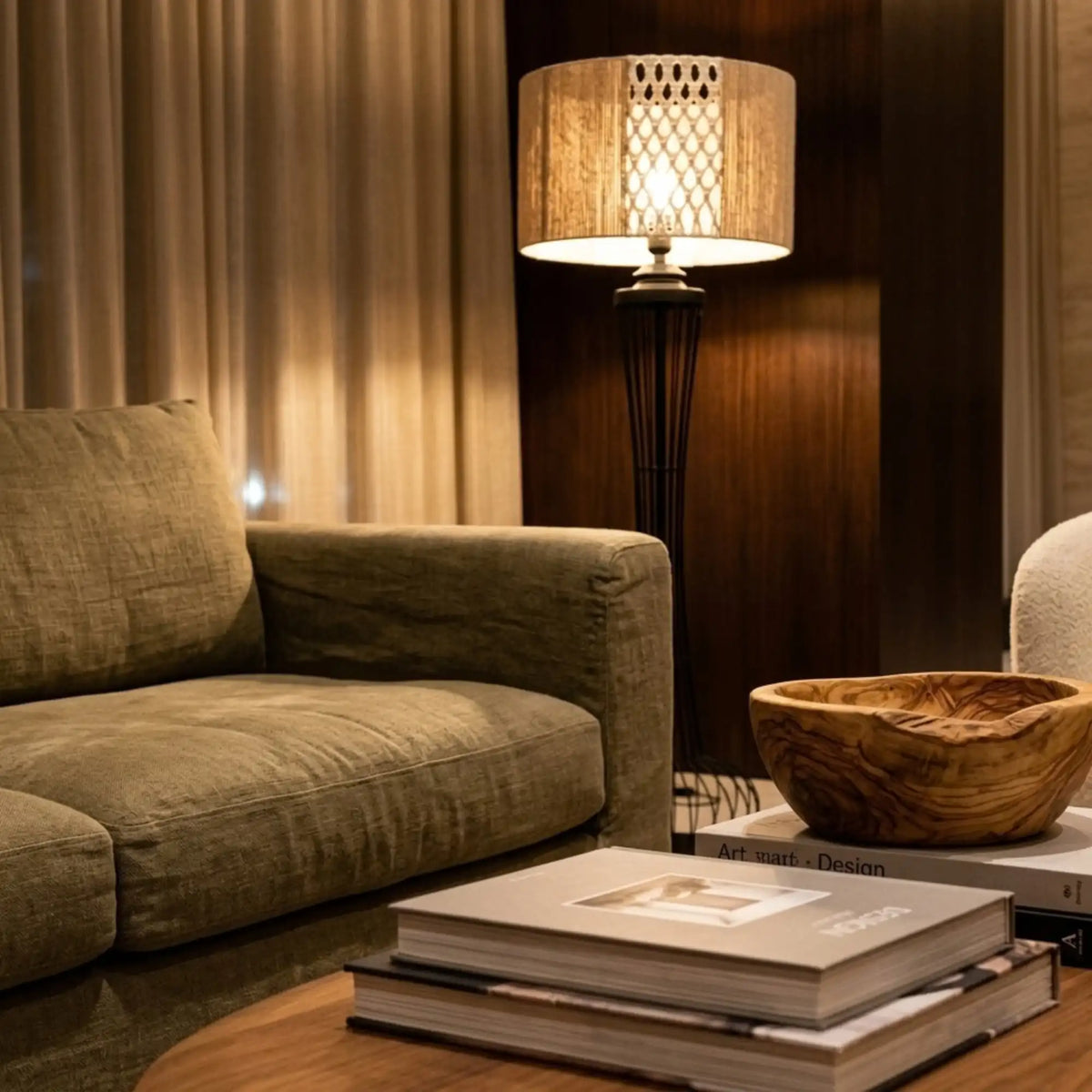 Lampe sur pied en jute et macramé allumée derrière un canapé vert kaki, près d'une table basse avec livres et coupe en bois