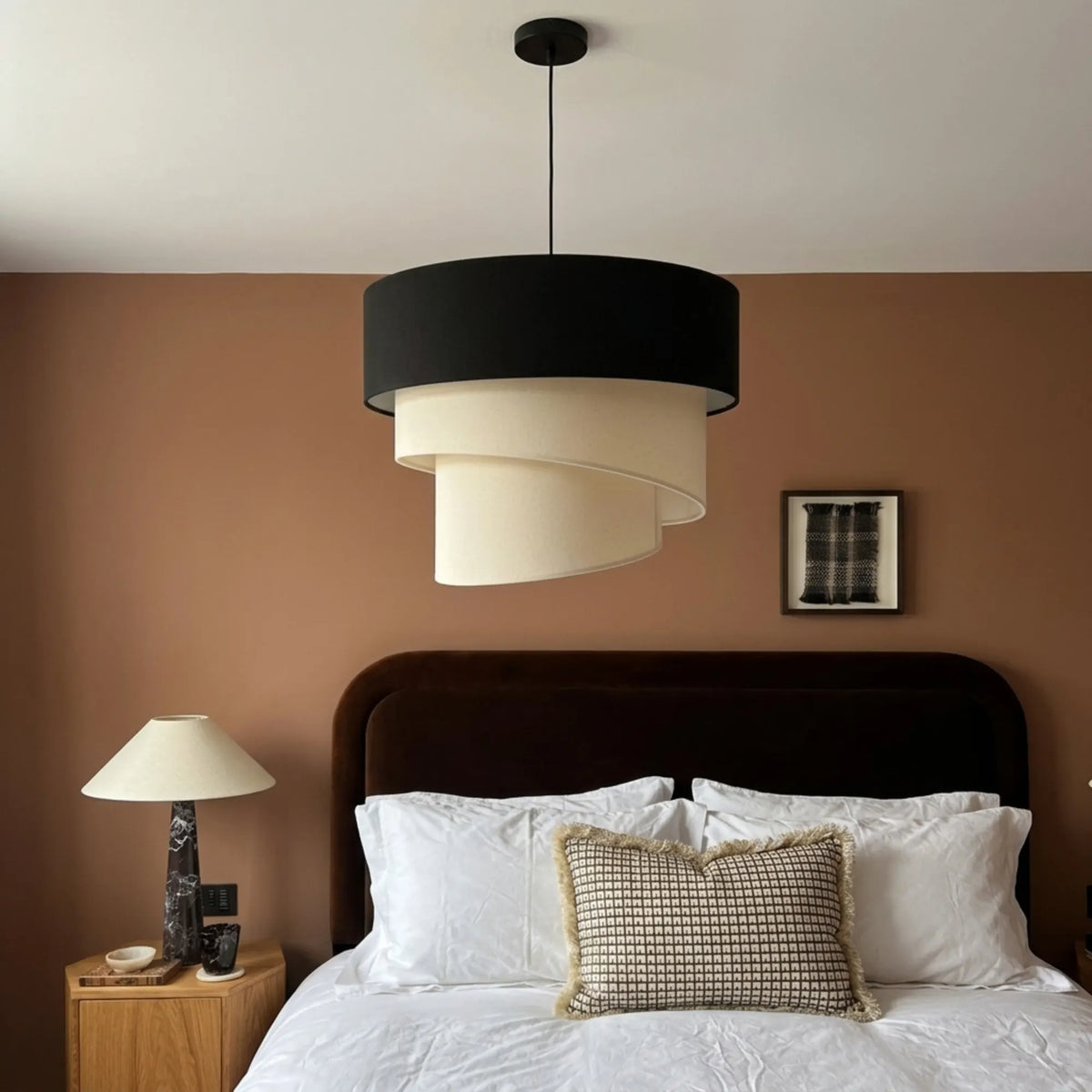 Suspension design noir et écru devant un mur terracotta, au-dessus d'un lit blanc